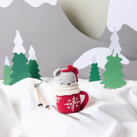 Crocheted gray cat with a red bow sitting inside a red mug decorated with white snowflakes. The cozy winter scene includes small wooden skis and ski poles on a white fabric surface, with a backdrop of stylized green pine trees and gray paper mountains. Perfect for Christmas décor, winter crafts, and DIY crochet ideas.