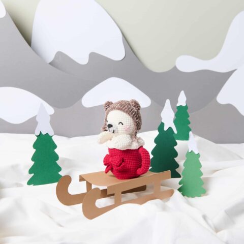 Eine gehäkelte Figur in einer Papierwelt auf einem Schlitten und in winterlicher Kulisse