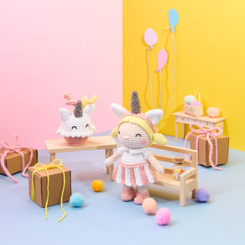 Bunte Amigurumi-Szene mit gehäkelten Einhornfiguren, Geschenken und Pompons vor pastellfarbenem Hintergrund – kreative Deko-Idee für DIY- und Häkelprojekte.